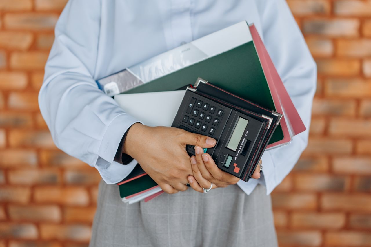 services-01 Close-up of a person holding a calculator and folders against a brick wall background.